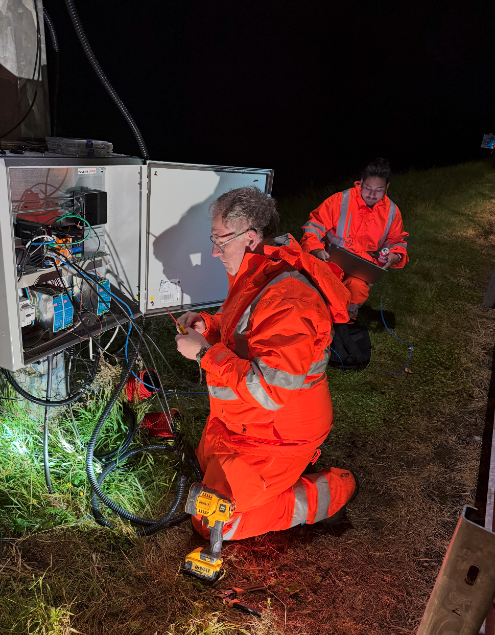 Two engineers in high-visibility clothing at night, wiring a roadside control cabinet in the field.