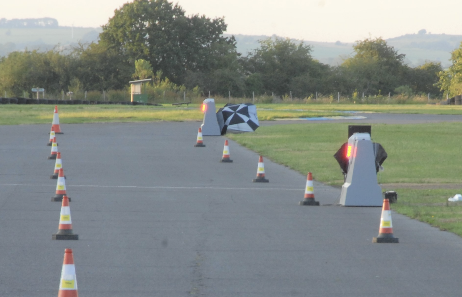 Roadside warning units with red lights illuminated, positioned among traffic cones on a test track at dusk.
